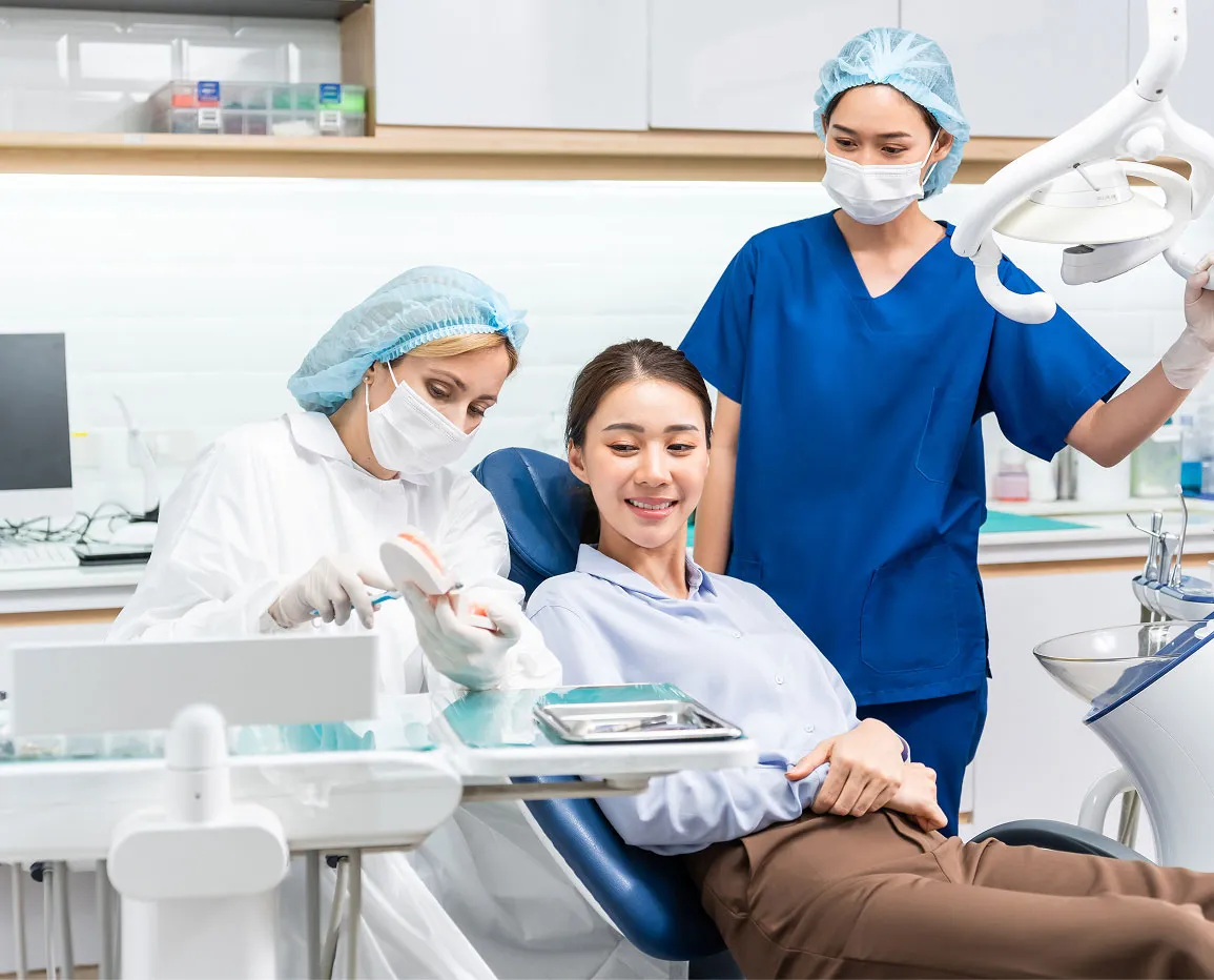Dentist in white coat and mask showing a dental model to a smiling female patient seated in a dental chair, with a dental assistant in blue scrubs adjusting overhead light.