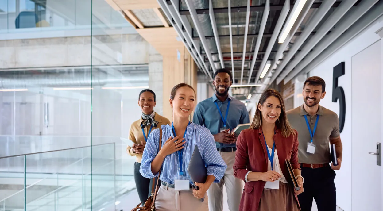 Five diverse business professionals walking together in a modern office hallway, smiling and holding folders and notebooks.