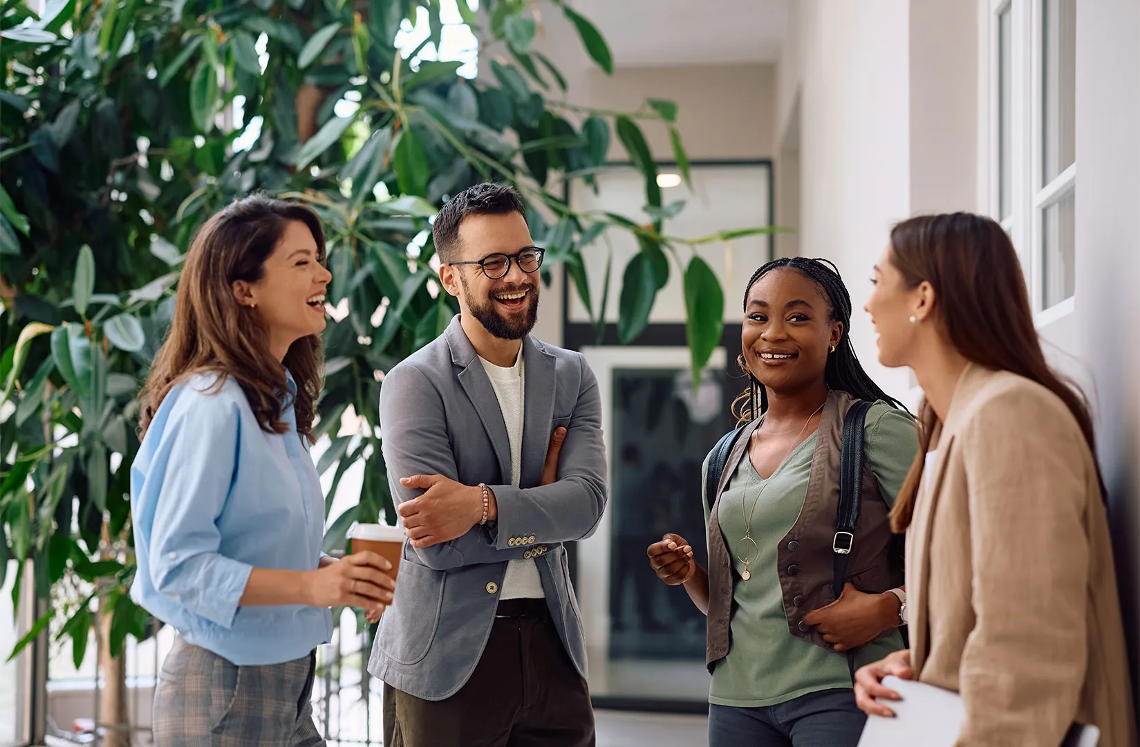 Four diverse colleagues smiling and chatting casually in a bright office hallway with large green plants.
