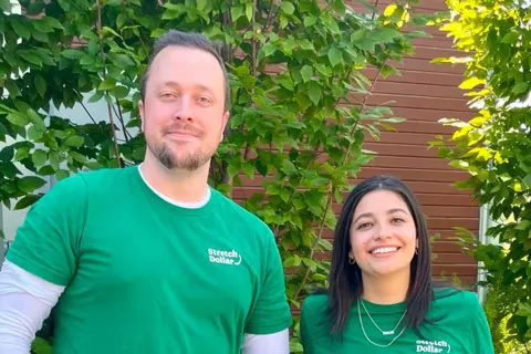 Co-founders of StretchDollar, Marshall and Kaiza, wearing StretchDollar shirts standing outdoors with green foliage and wooden wall behind.