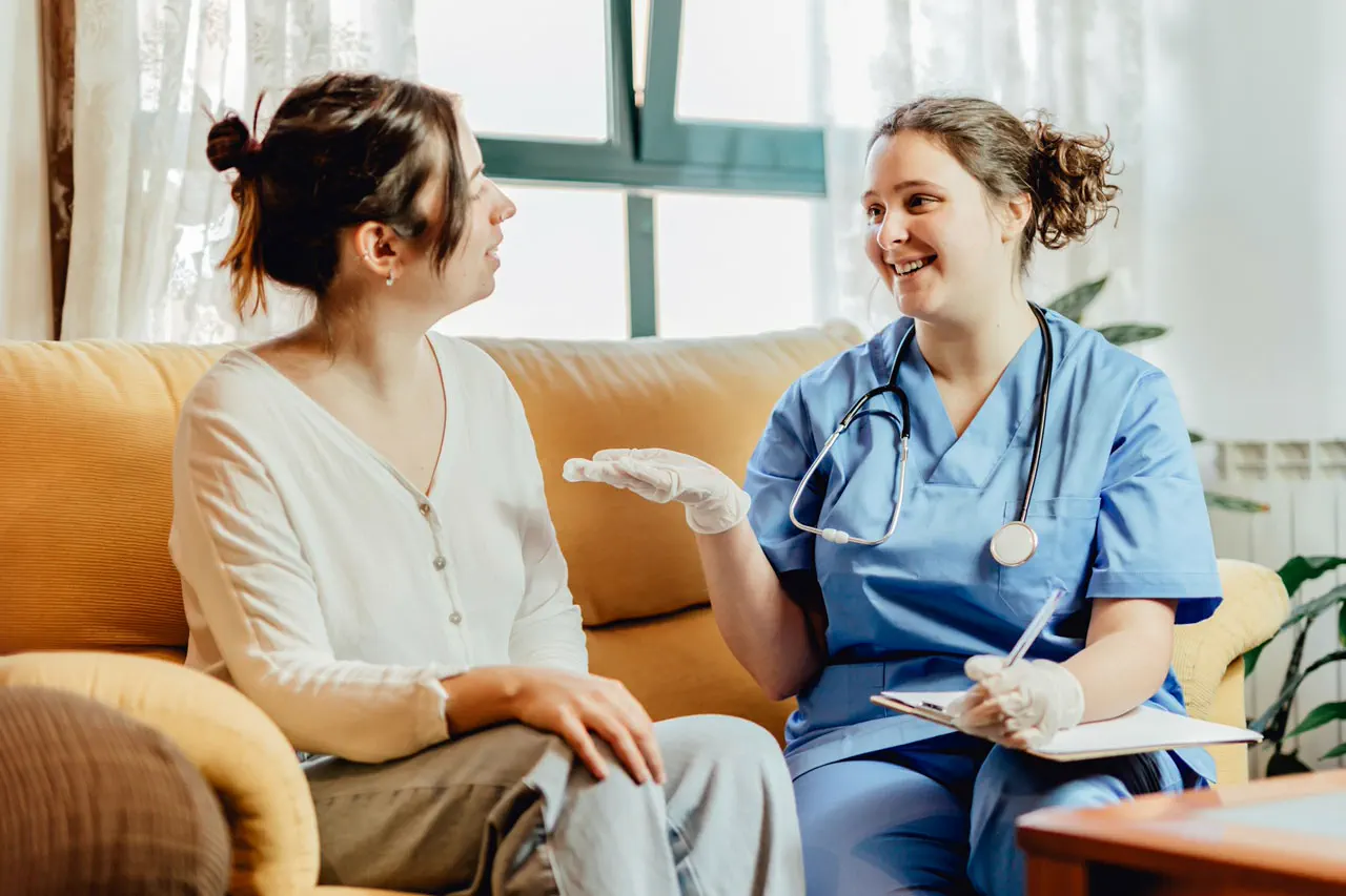 Female healthcare worker in scrubs talking to woman sitting on a yellow couch in a bright room.