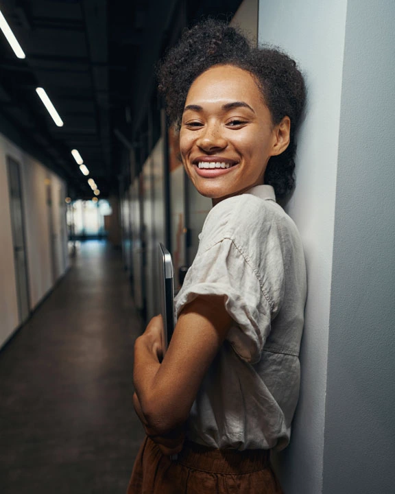 Smiling young woman leaning against a wall in a hallway holding a laptop.