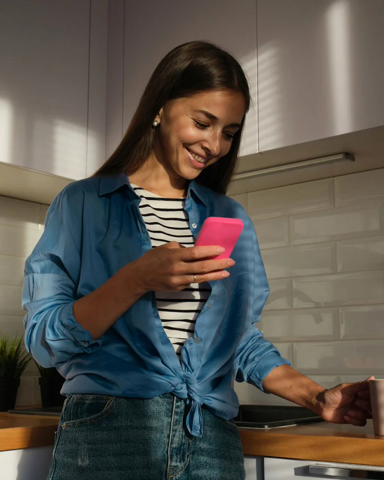Smiling woman in blue shirt looks at phone while holding a cup in a kitchen with wooden counters.