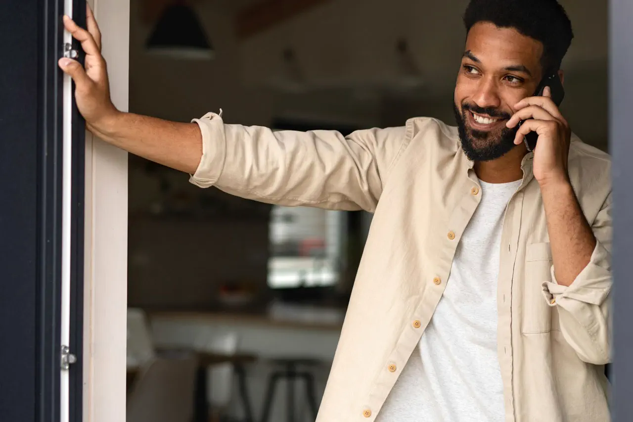 Smiling man in beige shirt talking on phone, leaning on doorframe indoors with blurred background.