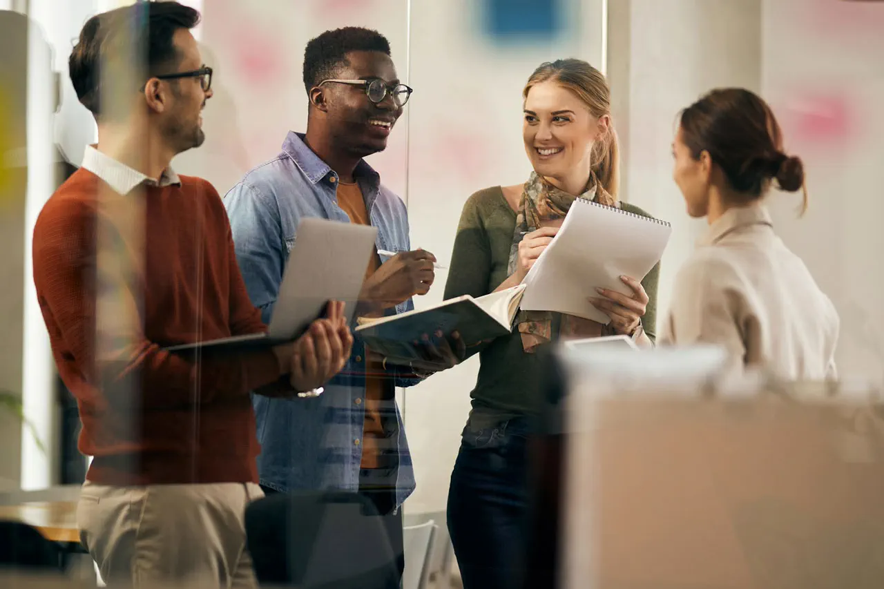 Four diverse colleagues smiling and discussing notes in a modern office meeting.