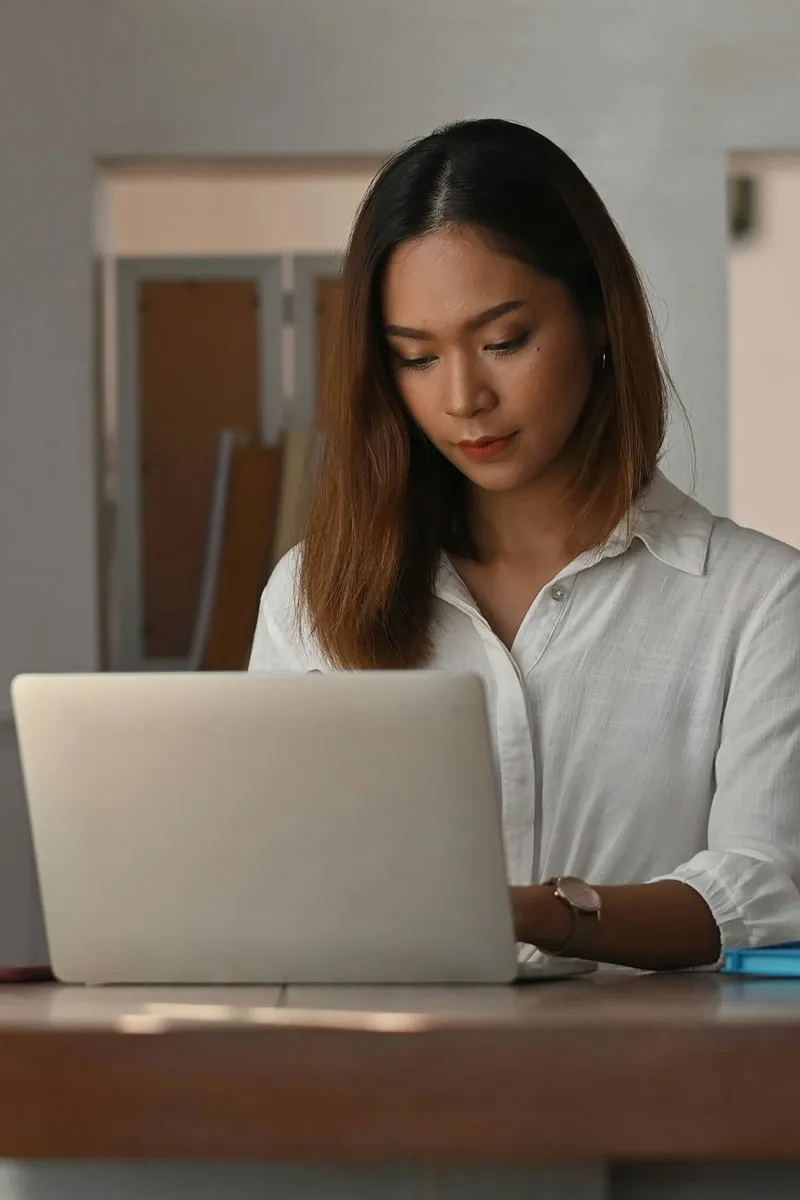Woman with shoulder-length hair working on a laptop at a wooden desk in a neutral-toned room.