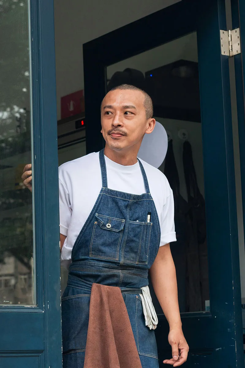 Man in a white shirt and denim apron standing at a blue door, looking to the side.