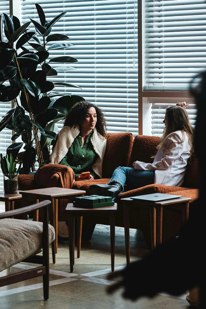 Two women seated on a brown sofa talking in a room with large blinds and plants.
