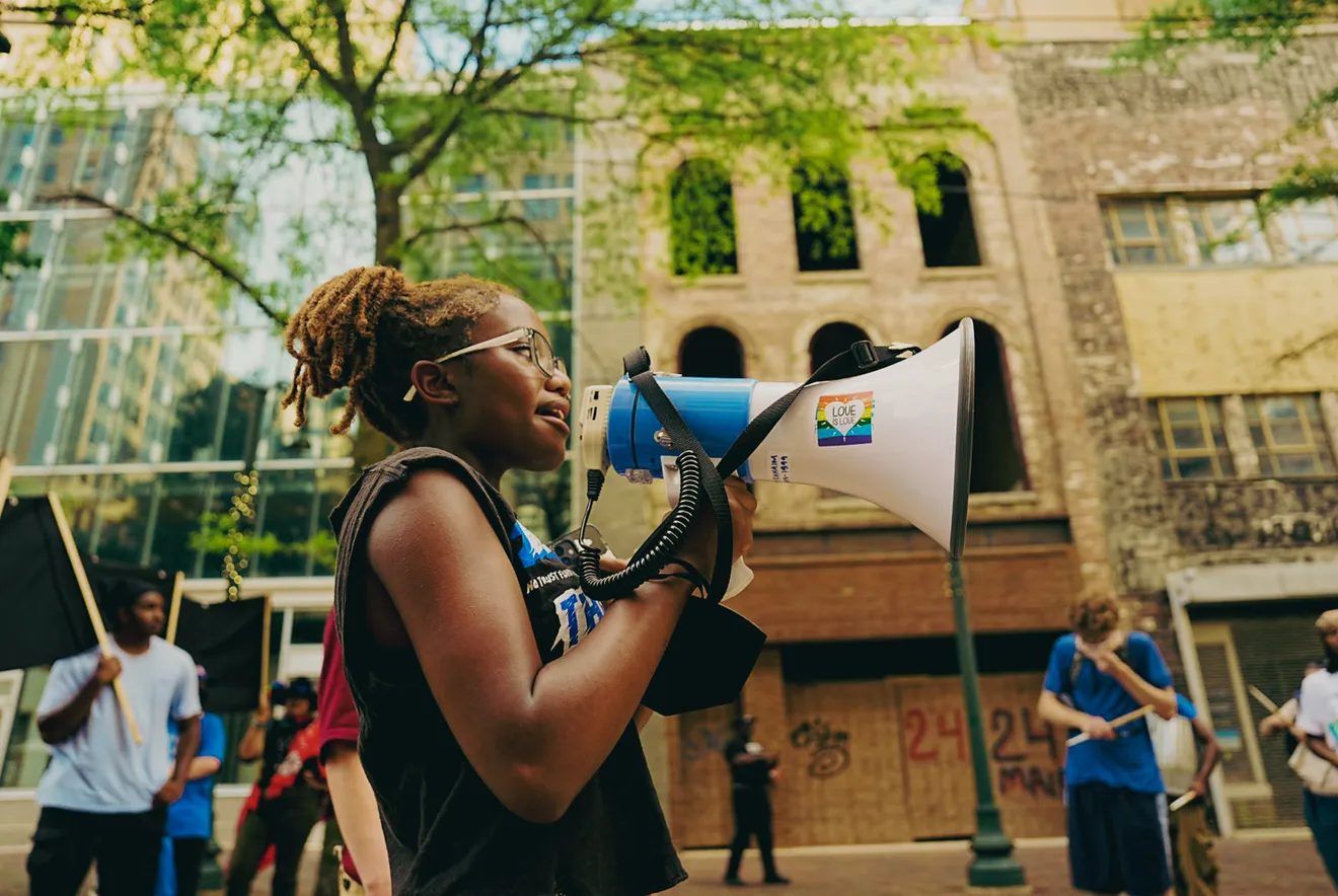 A young protester speaking into a megaphone during a street demonstration, with other participants and buildings in the background.