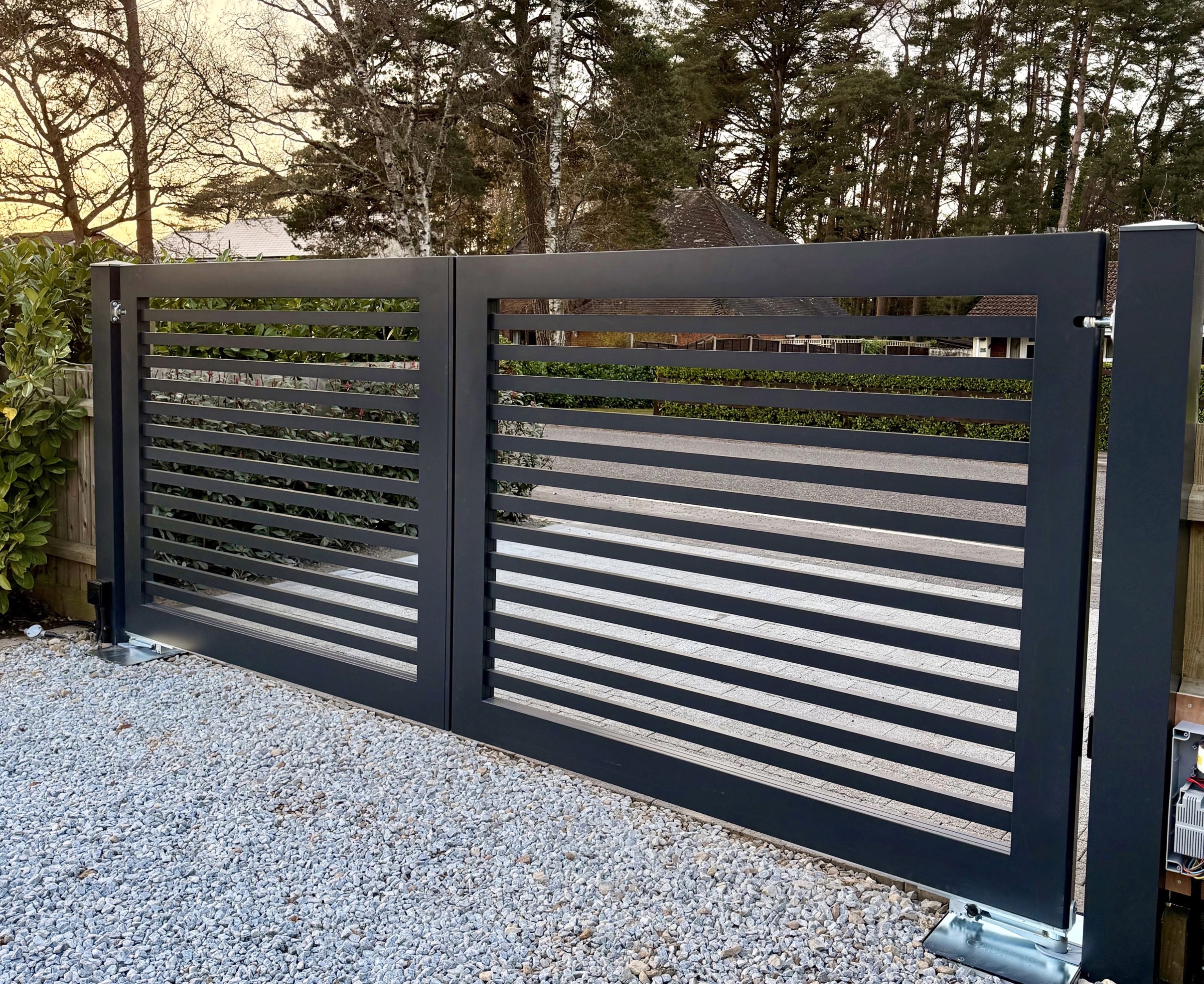 Modern black metal driveway gate with horizontal slats, set on a gravel surface with trees and bushes in the background.