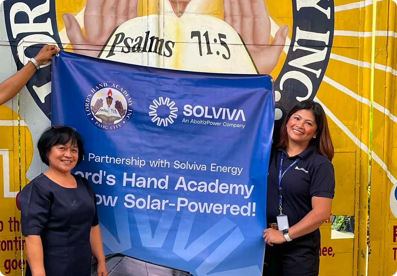 Two women holding a Solviva Energy banner celebrating Lord's Hand Academy going solar-powered, in front of a mural background