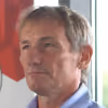 Portrait of an older man with short gray hair wearing a blue collared shirt, looking slightly to the side.