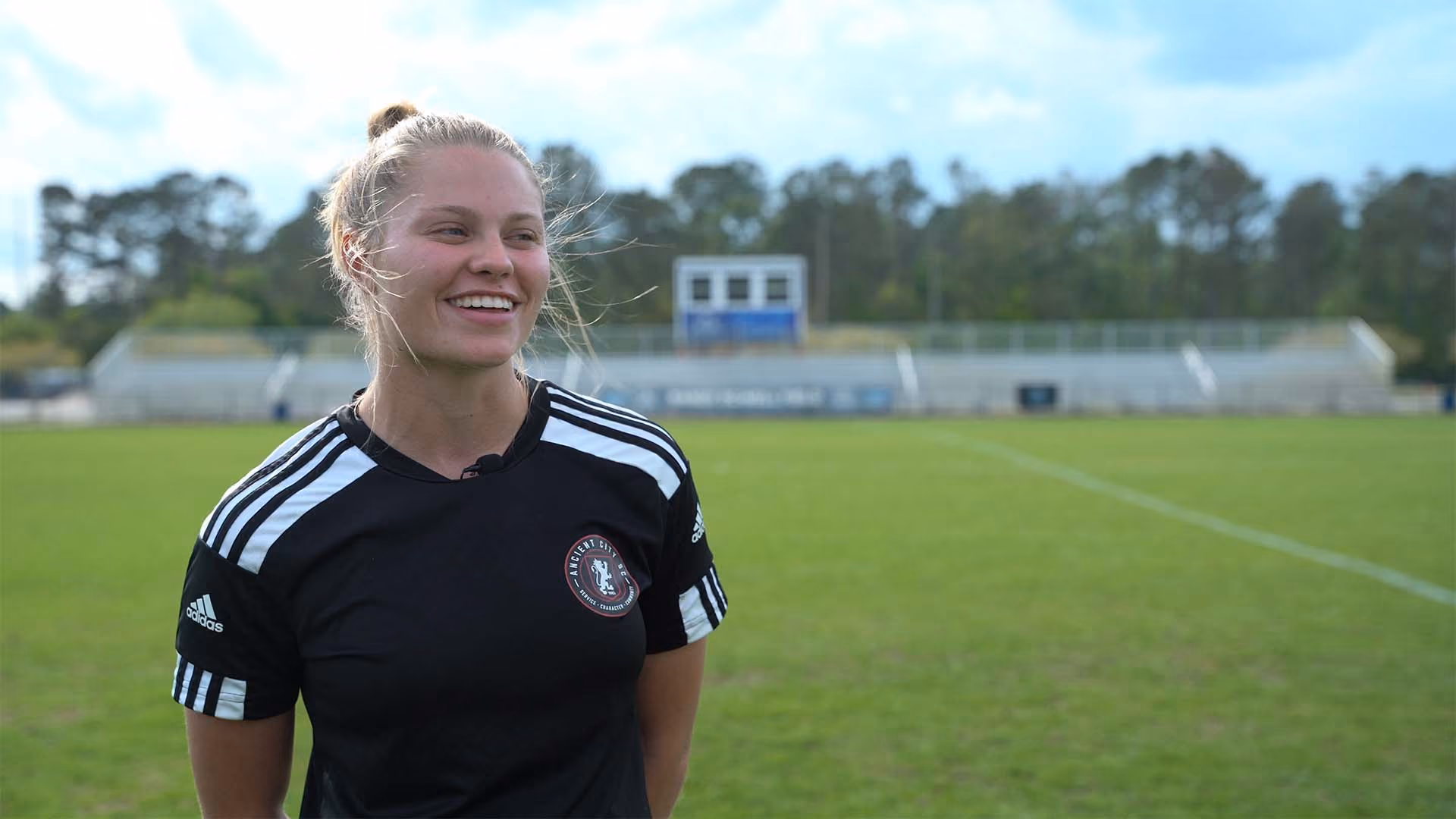 Smiling woman with blond hair in a black soccer jersey standing on a green soccer field with bleachers in the background.