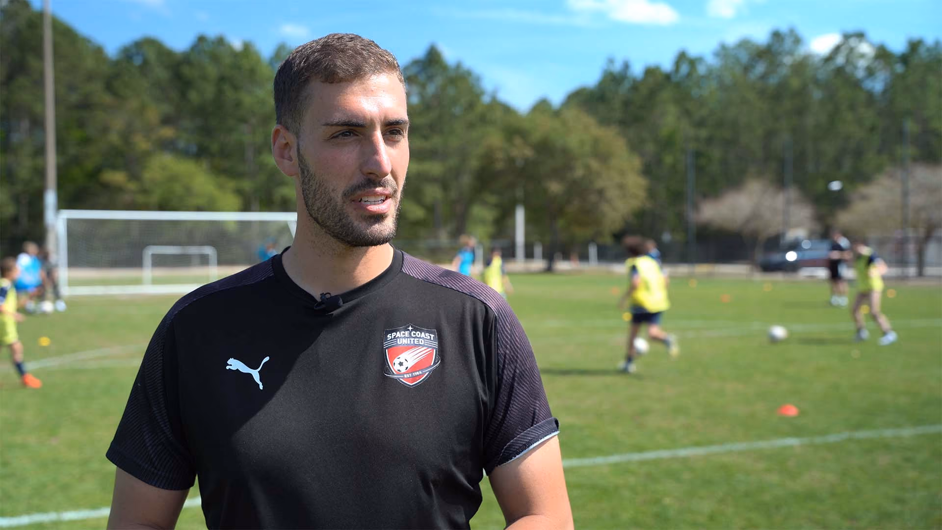 Man in a black Space Coast United soccer shirt standing on a soccer field with players practicing in the background.
