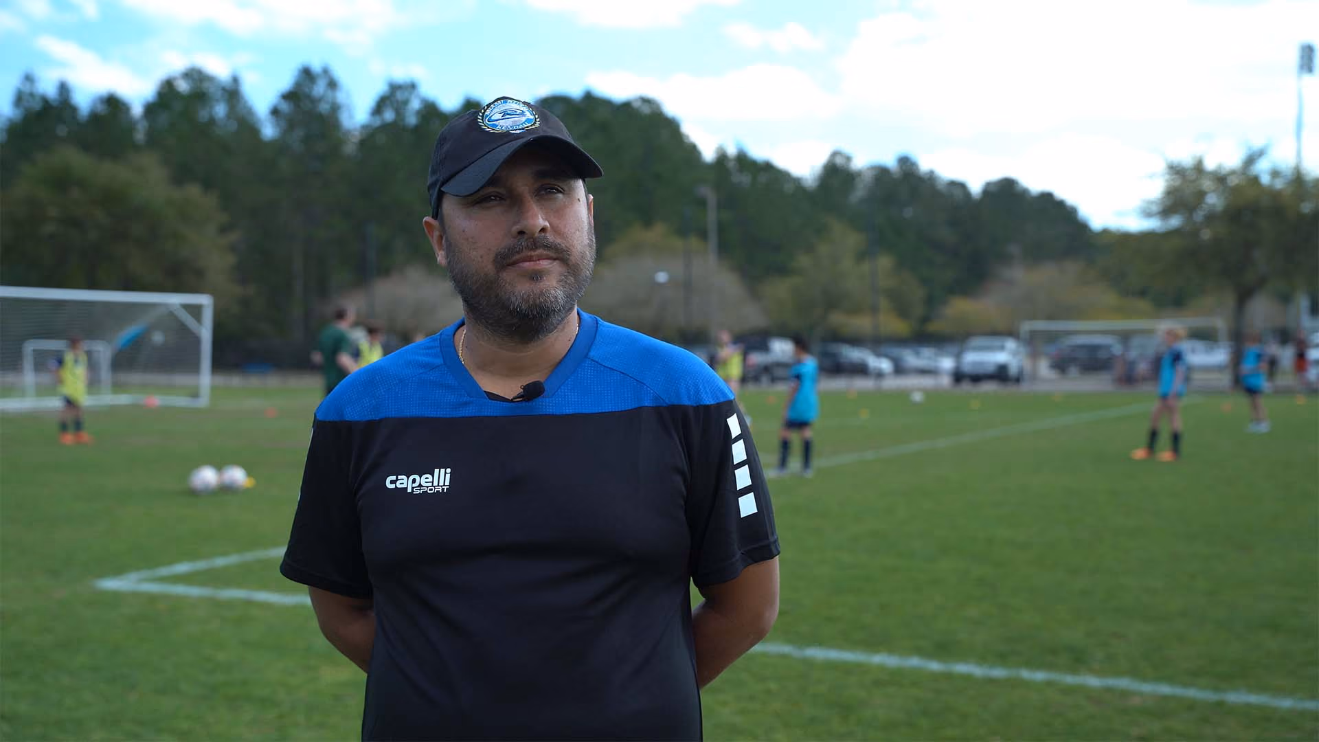 Man wearing a black and blue sports shirt and cap standing on a soccer field with players and goals in the background.