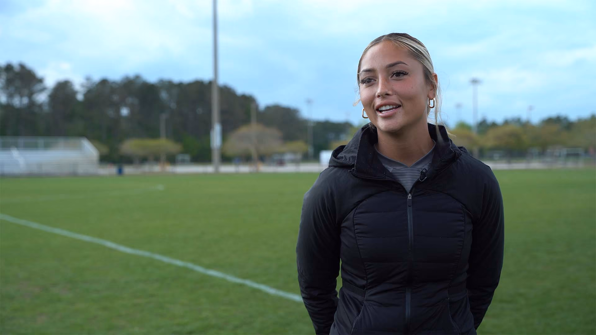 Young woman with blond hair in a black jacket standing on a sports field with trees and bleachers in the background.