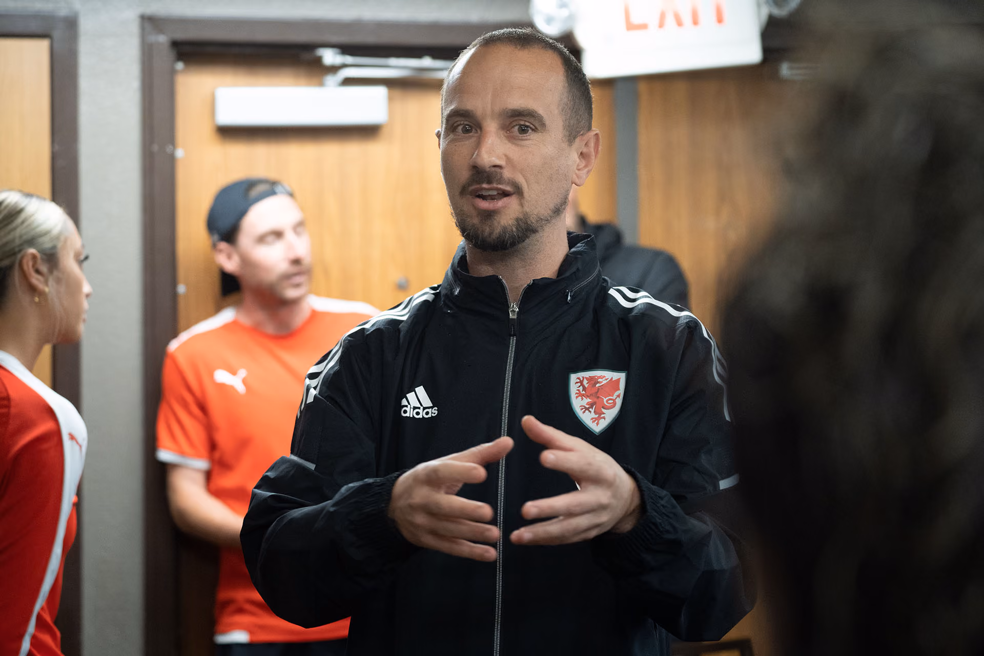 Man in black Adidas jacket with Wales emblem speaking and gesturing with hands in a room with other people.