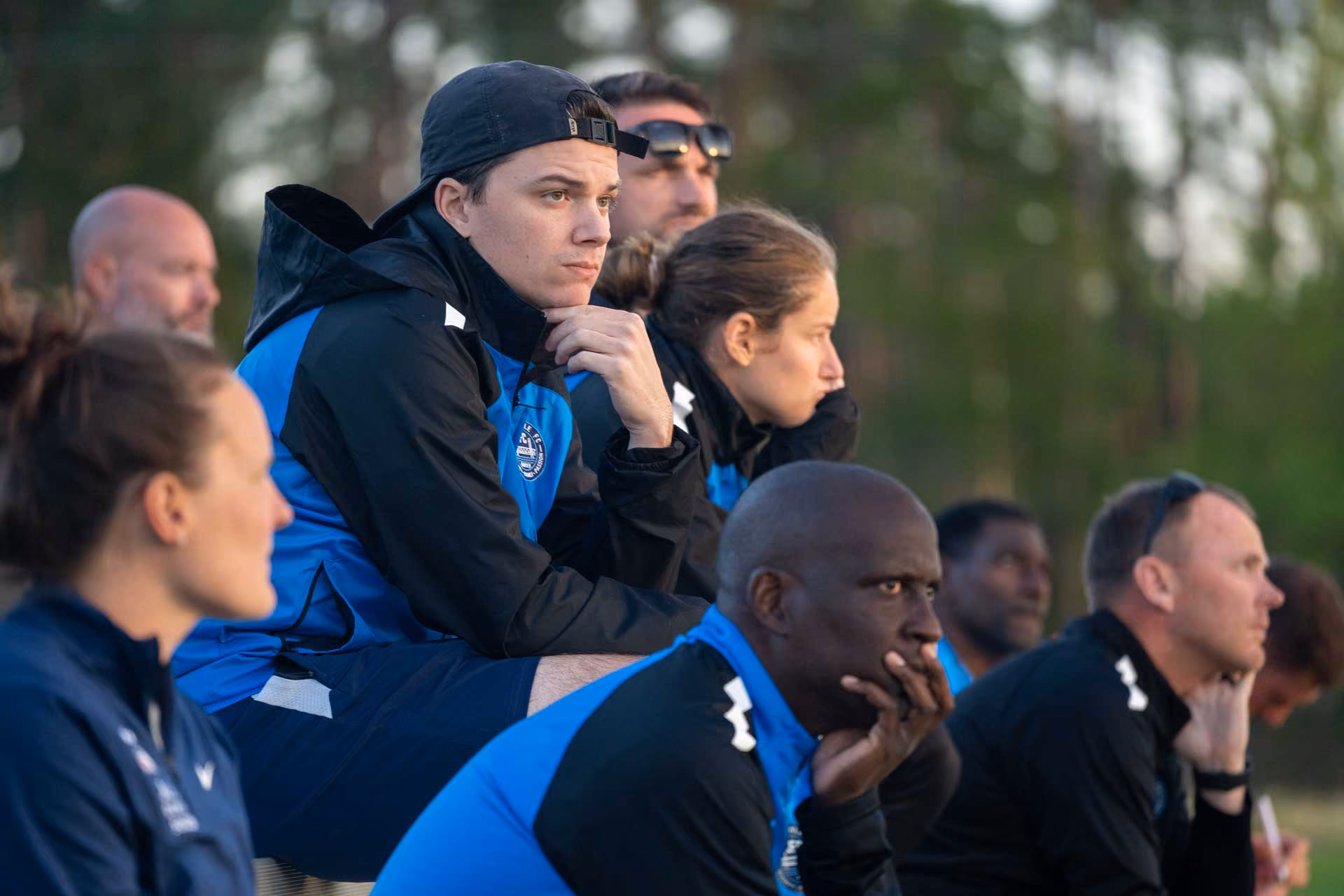 A group of sports coaches and players sitting on a bench outdoors, attentively watching something off-camera.