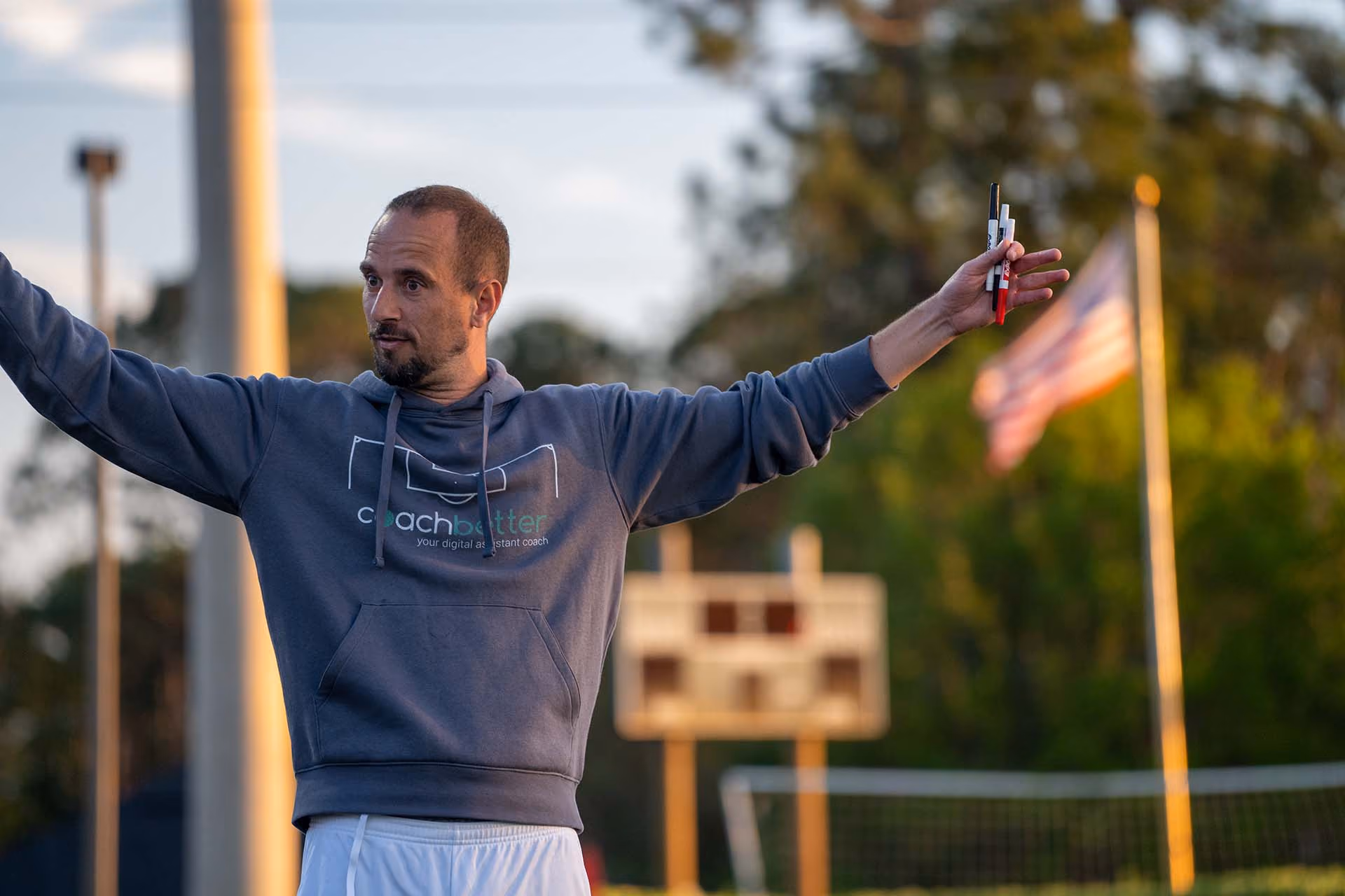 Man in a hoodie with 'coachbetter' logo holding markers, standing outdoors with a scoreboard and an American flag in the background.