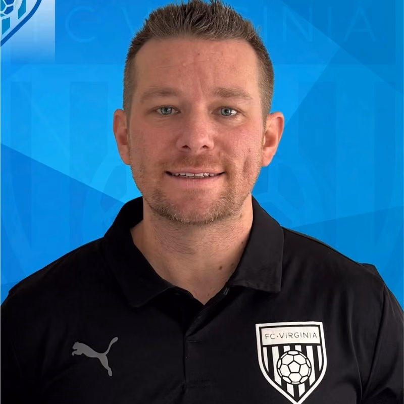 Man with short brown hair and a trimmed beard wearing a black FC Virginia shirt with Puma logo against a blue background.