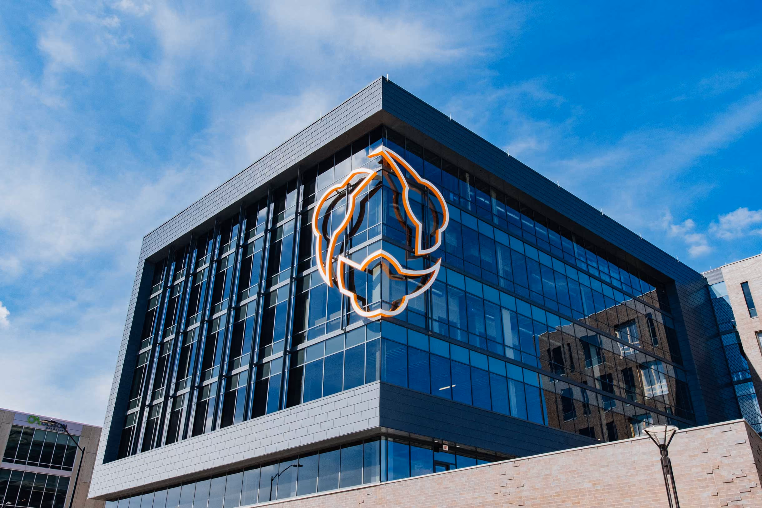 Modern office building with large glass windows featuring an orange and white outlined logo on the corner under a partly cloudy blue sky.
