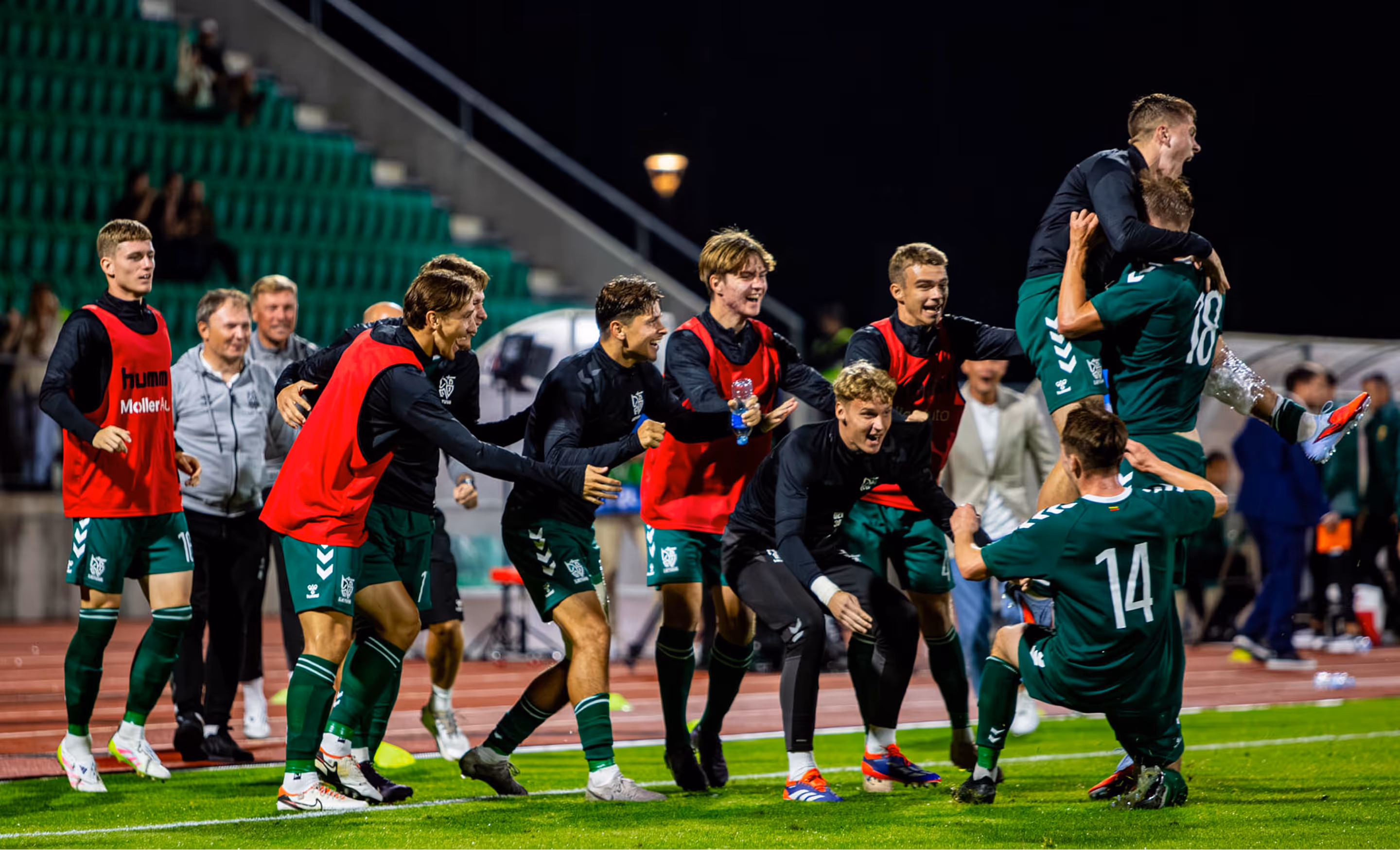 Soccer players celebrating on a field at night, with some wearing red training vests and others in green uniforms.