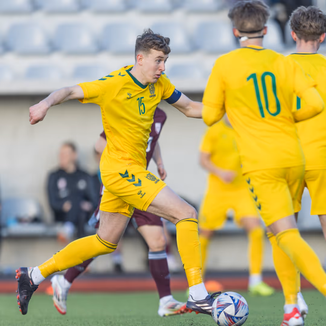 Soccer player in yellow jersey number 15 kicking a soccer ball during a match with players in yellow and maroon in the background.
