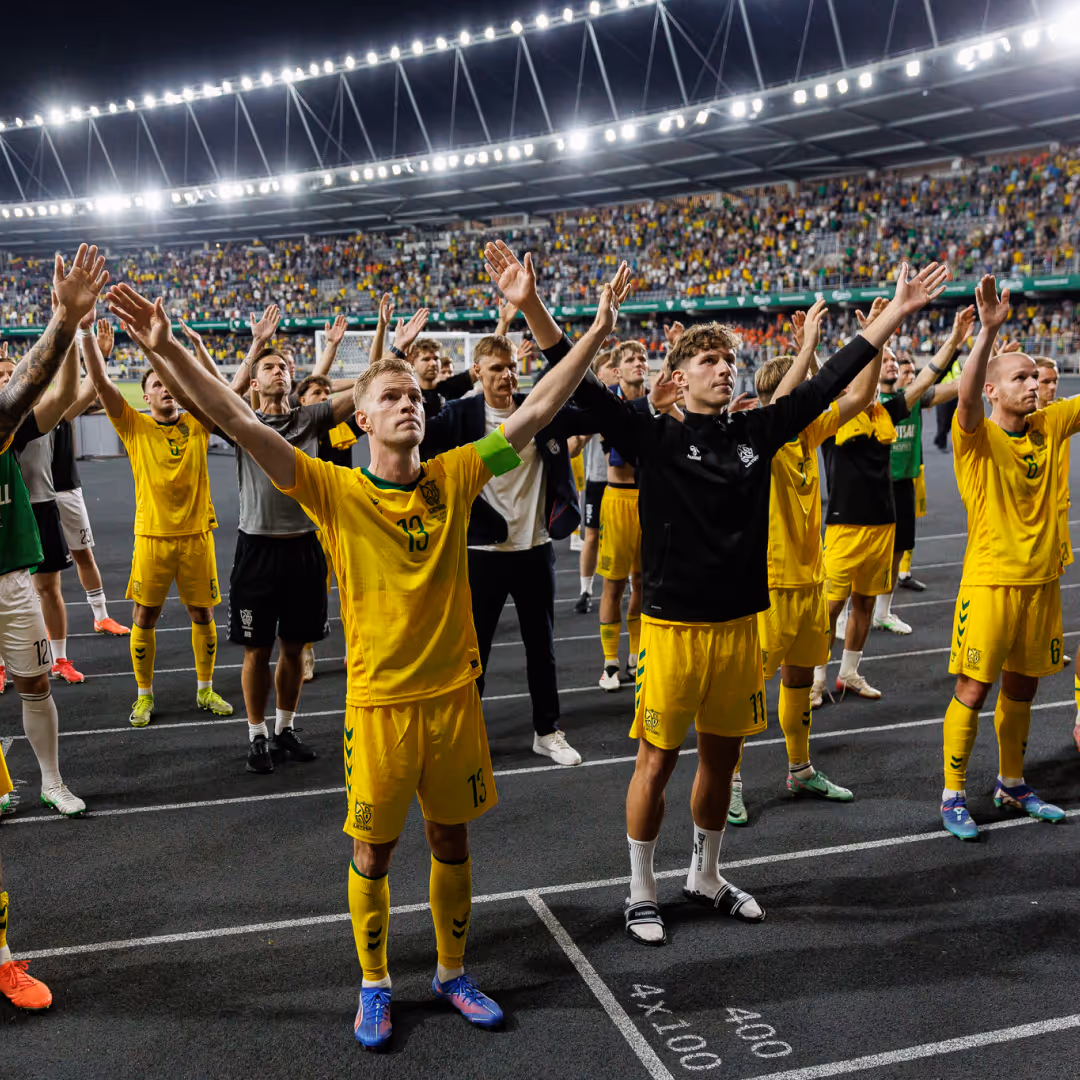 Soccer players in yellow uniforms standing on the field with arms raised, facing crowd in a brightly lit stadium.