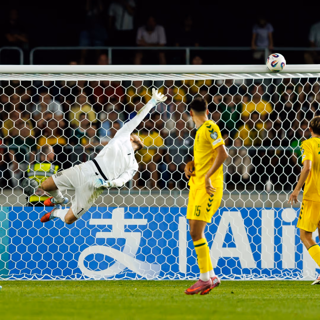 Soccer goalkeeper in white diving to try and save a ball heading toward the goal with players in yellow watching.