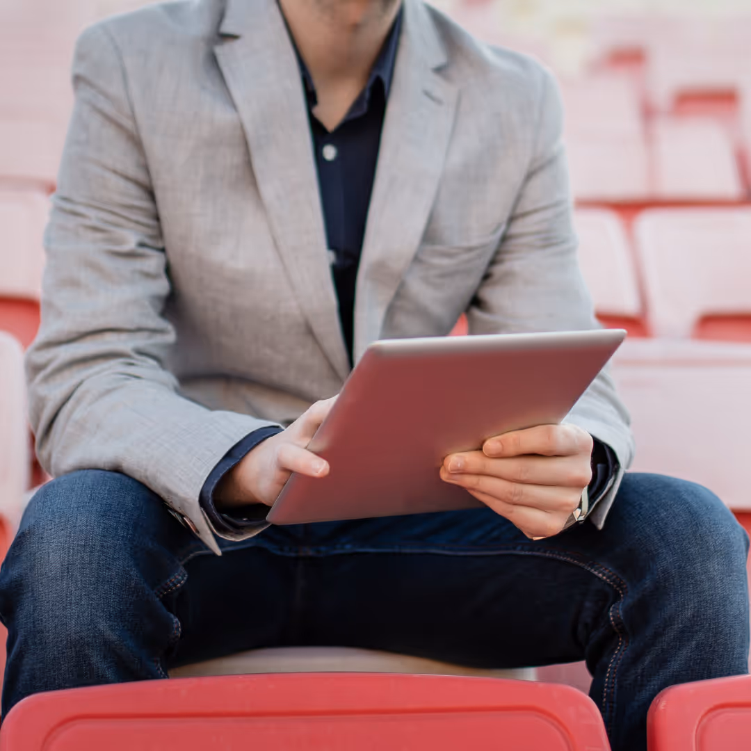 Man in gray blazer and jeans sitting on red stadium seats while holding a tablet.