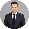 Man in a black suit and tie with a white shirt standing against a light gray background.