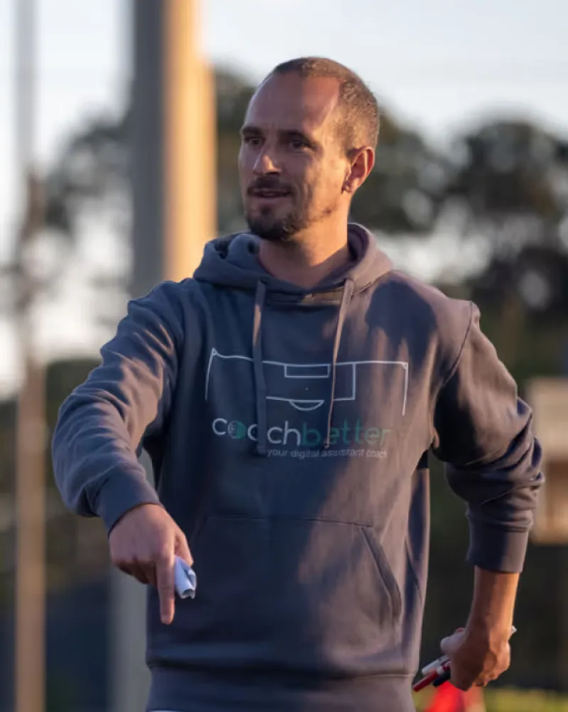 Coach in a grey Coachbetter hoodie pointing down while giving instructions on a pitch