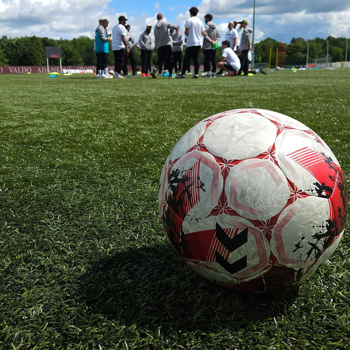 Football on a grass pitch with a group of players gathered in the background