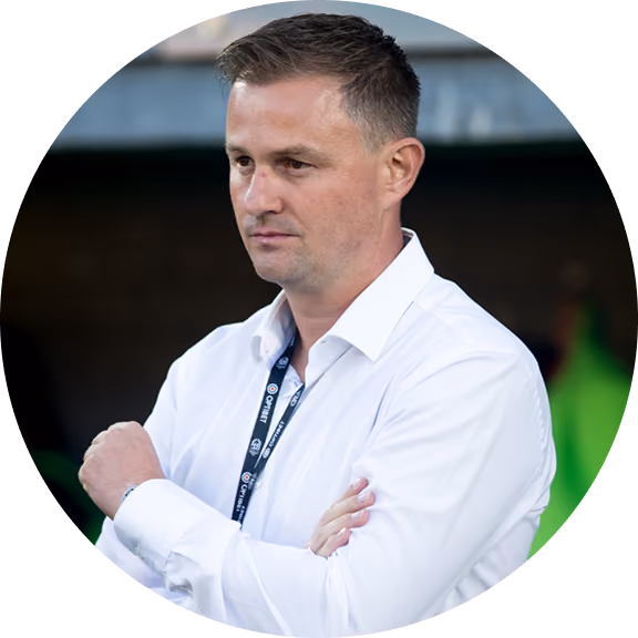 Man in a white shirt with a lanyard standing with arms crossed in a stadium