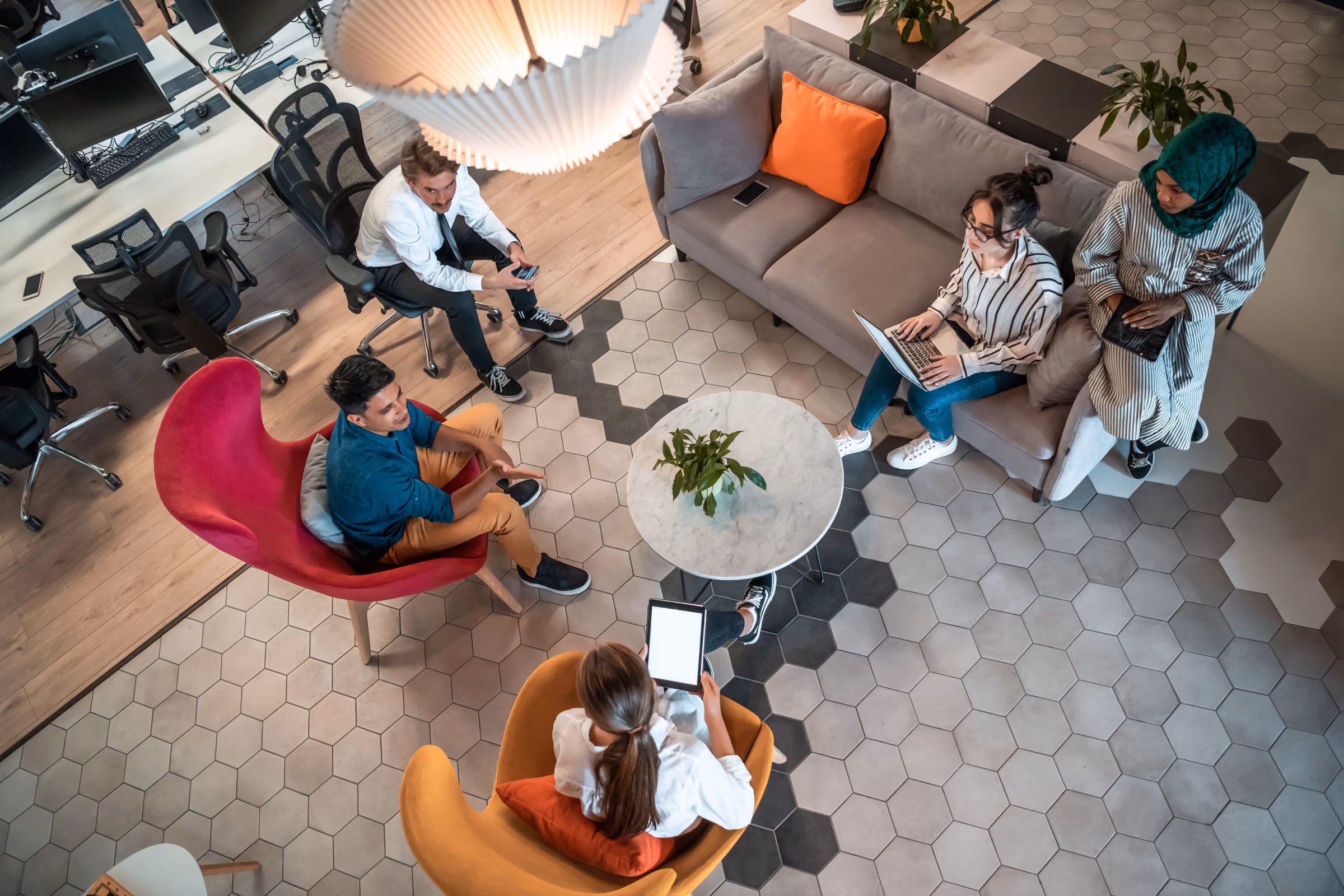 Five diverse young adults in a modern office lounge collaborating around a round table, with some using laptops and tablets while seated on a gray couch and colorful chairs.
