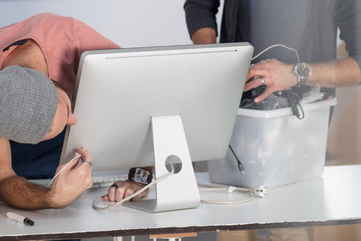 Two men working on setting up or fixing an all-in-one computer on a white table, one bending down connecting cables, the other holding a white plastic container with cables and devices.