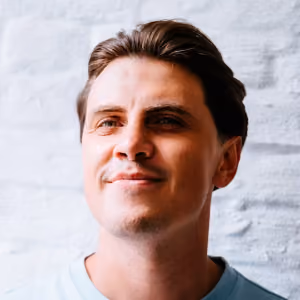 Close-up of a smiling man with short dark hair wearing a light blue shirt standing in front of a white brick wall.