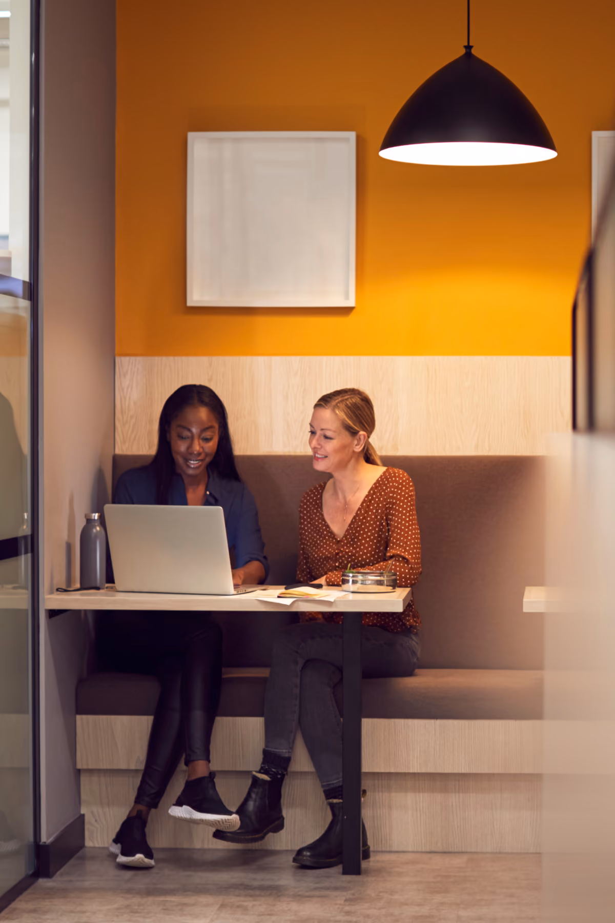 Due donne sedute su una panca in un ufficio moderno con parete arancione, che lavorano insieme su un laptop posto su un tavolo con libri e una borraccia.