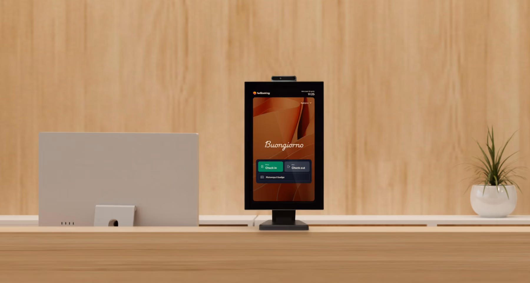 Dispositivo digitale su scrivania in legno, con schermo che mostra un'interfaccia di check-in e check-out in italiano con la scritta Buongiorno, accanto a un monitor grigio e una piccola pianta in vaso bianco.