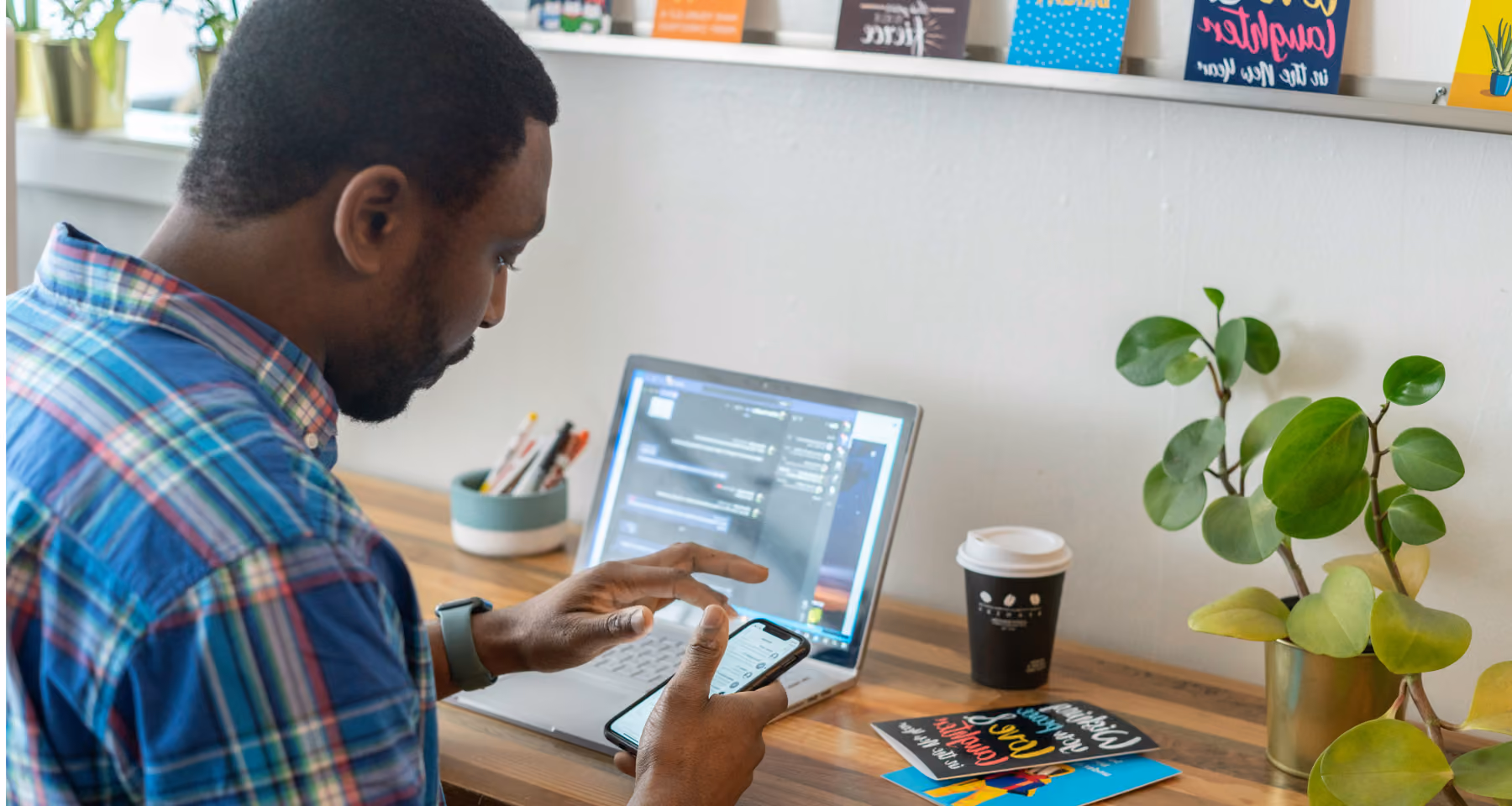 Uomo seduto a una scrivania che usa uno smartphone con un laptop aperto davanti a sé, circondato da una pianta, alcuni libri e una tazza di caffè sul tavolo.