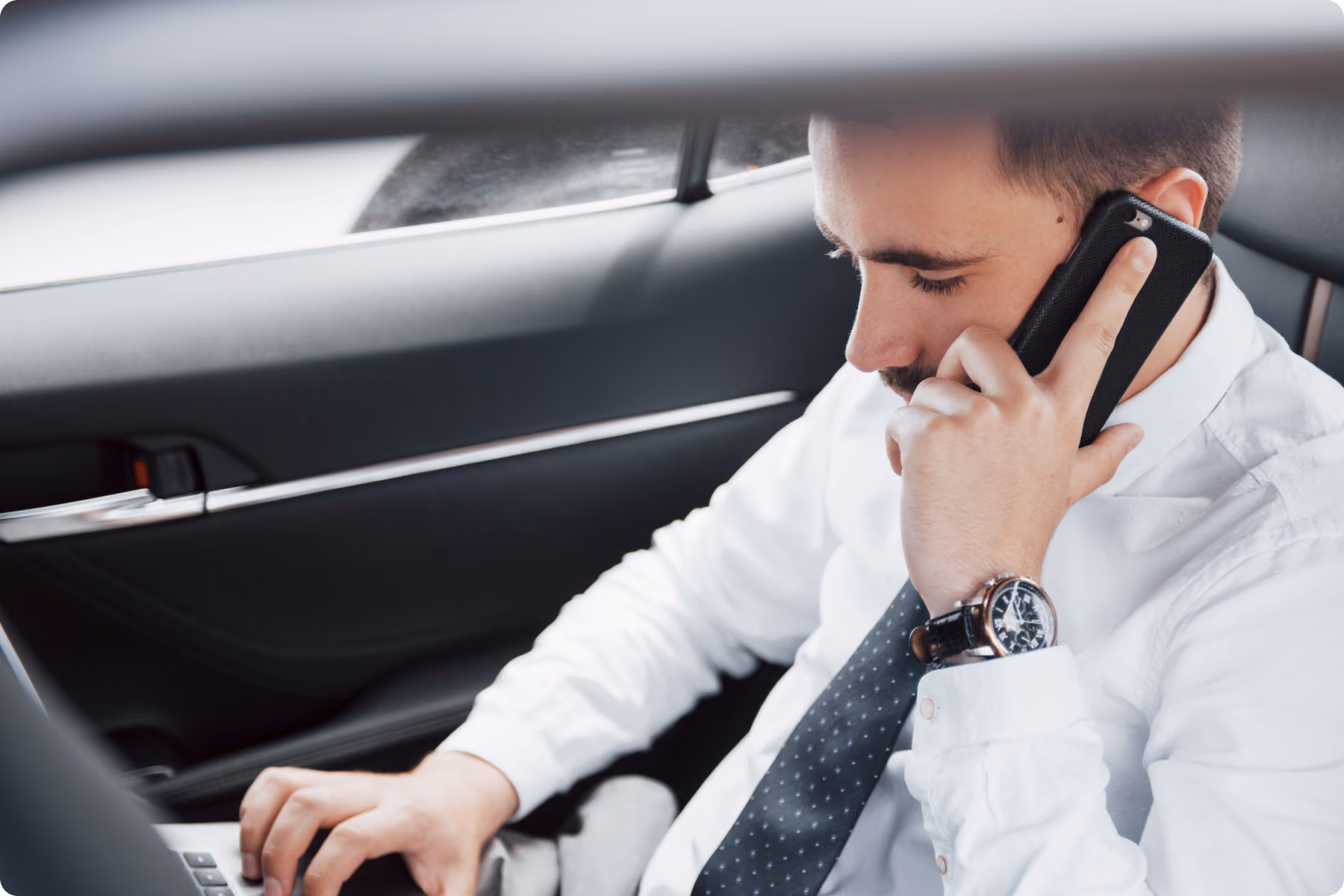 Uomo d'affari seduto in macchina che parla al cellulare mentre usa un computer portatile.