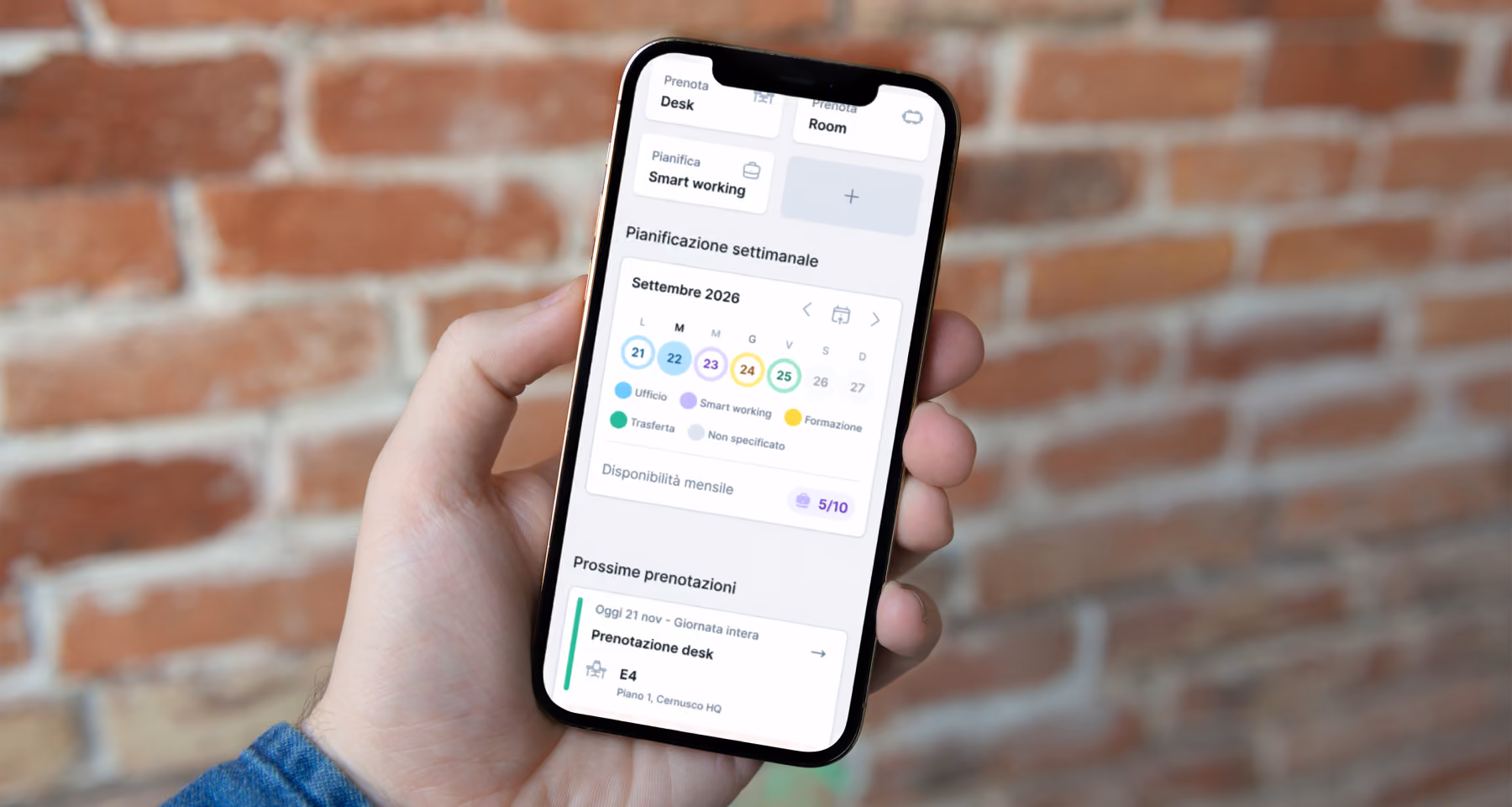 Mano che tiene uno smartphone con schermo che mostra un'app di pianificazione settimanale per settembre 2026, con calendario colorato per Ufficio, Smart working, Formazione, Trasferta e Non specificato e informazioni su disponibilità mensile e prenotazioni desk.