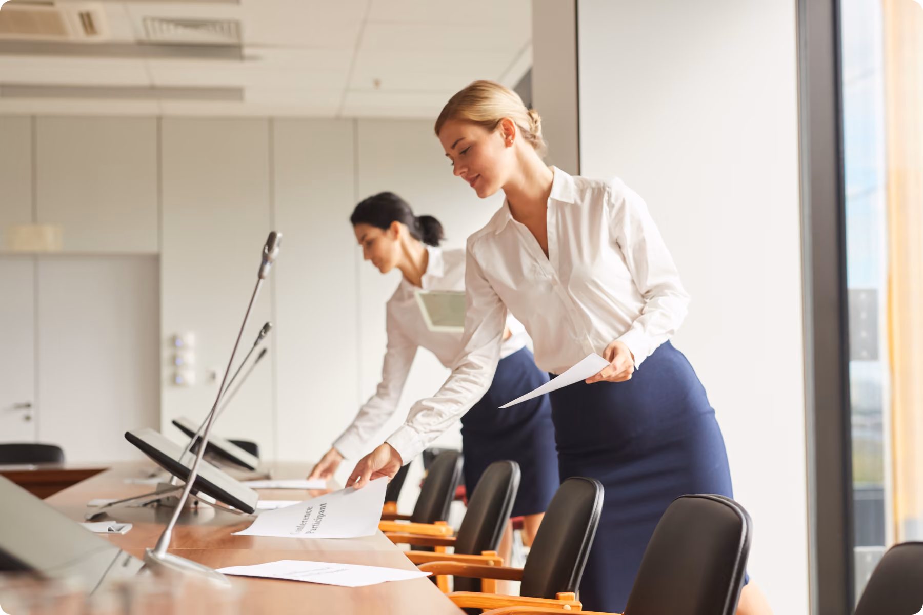 Due donne in abiti da ufficio, camicia bianca e gonna blu, che preparano documenti su un tavolo in una sala conferenze moderna con microfoni e sedie.