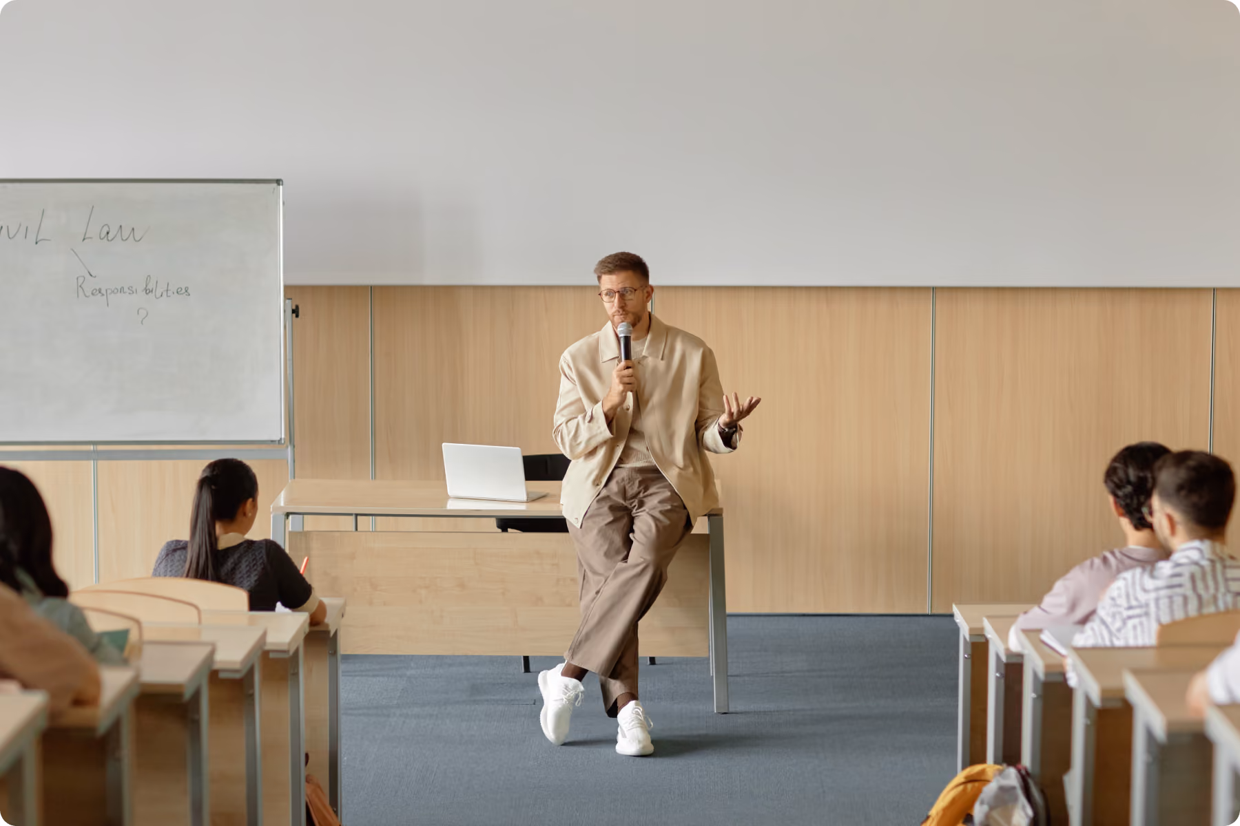 Insegnante uomo seduto sul tavolo di fronte agli studenti in aula, parla con microfono mentre sullo sfondo una lavagna con scritte sul diritto civile.