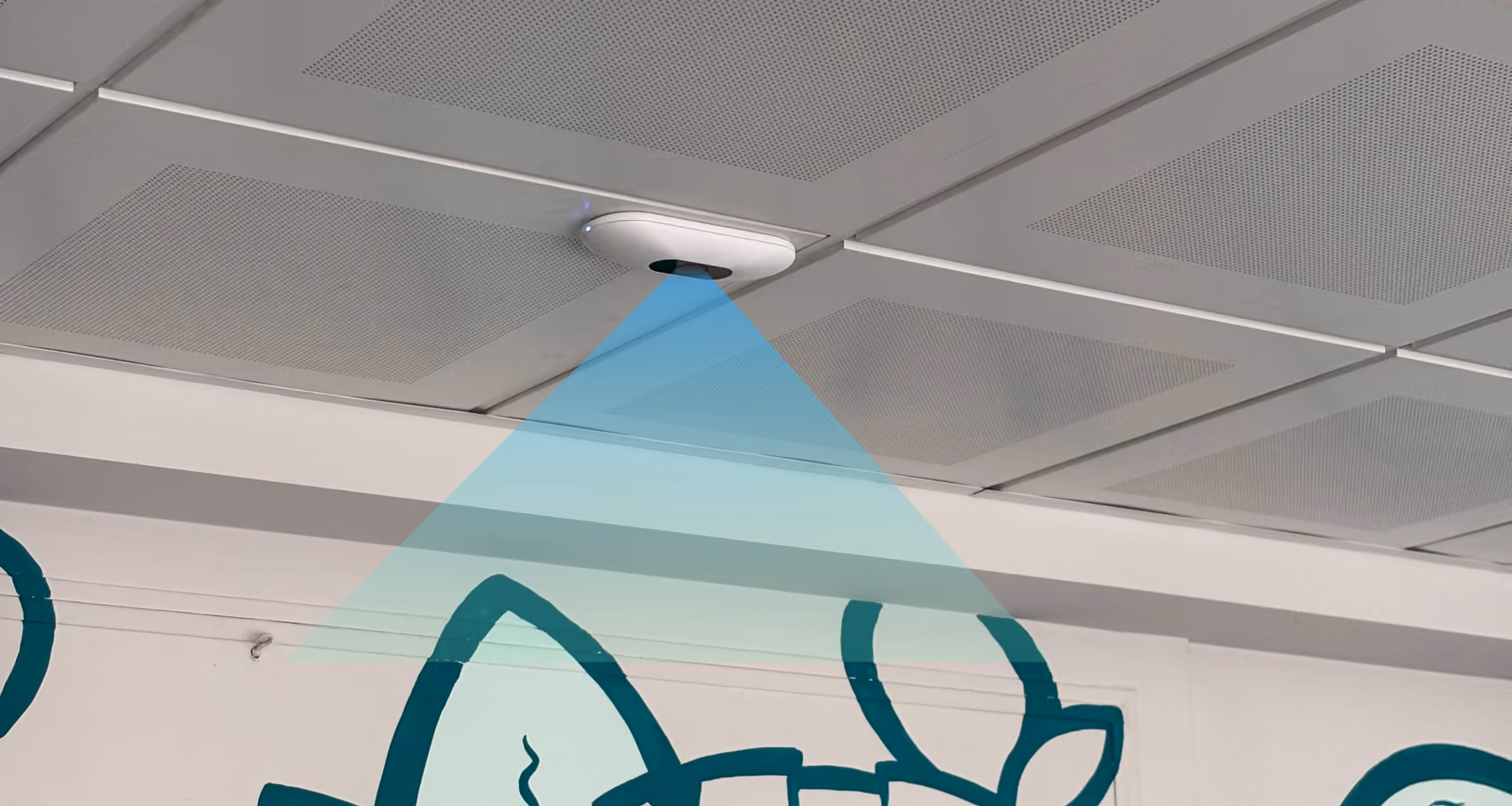 Sensore bianco montato sul soffitto di una stanza con pareti bianche decorate da pitture murali verdi; un raggio azzurro indica l'area sorvegliata dal sensore.