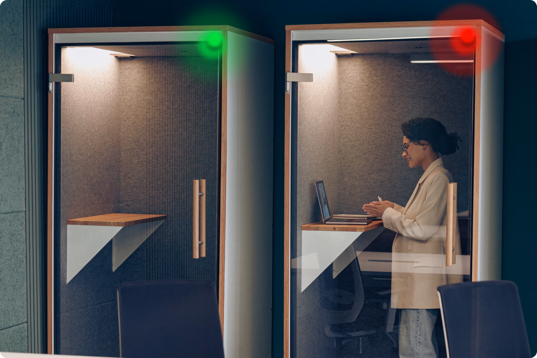 Donna d'affari che parla in videochiamata in una cabina acustica da ufficio ben illuminata con luce interna, mentre nell'altra cabina è vuota con una luce verde intermittente.