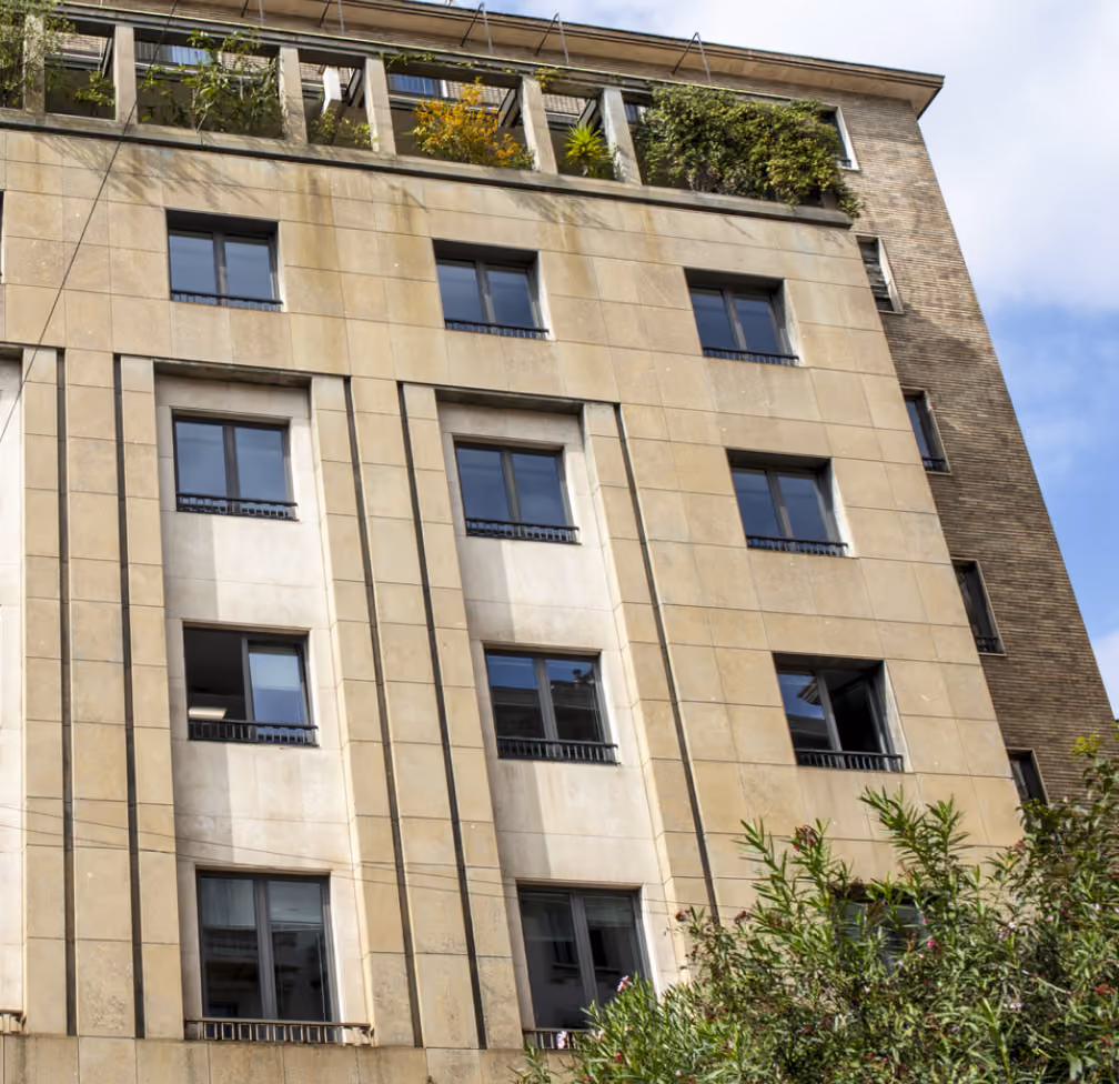 Facciata di un edificio moderno con pareti beige e numerose finestre rettangolari; sul tetto ci sono piante verdi in vaso e al centro in basso si intravede una pianta verdi e fiori.