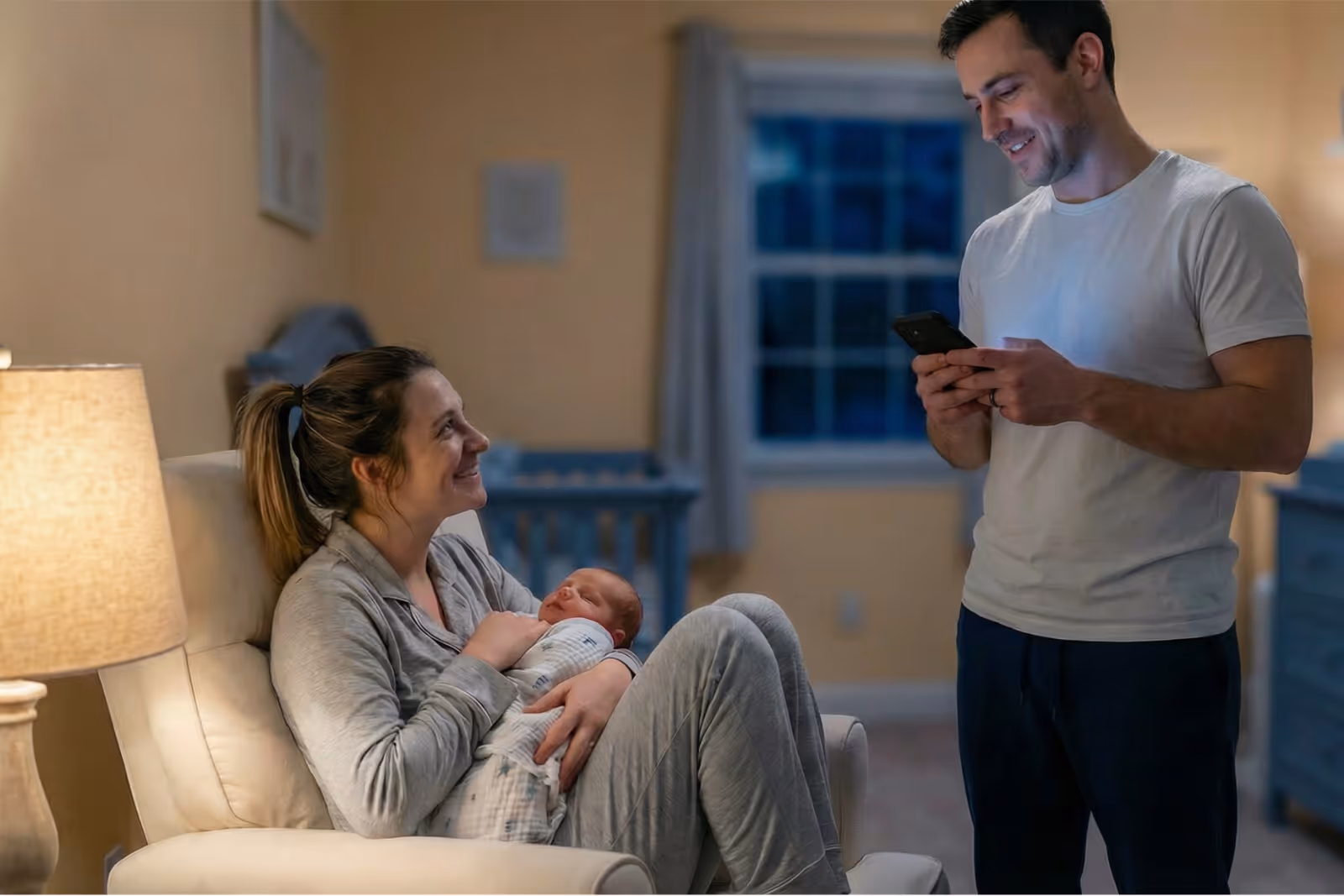 Smiling woman in gray pajamas cradling a sleeping newborn in a cozy nursery while a man standing nearby looks at his phone.