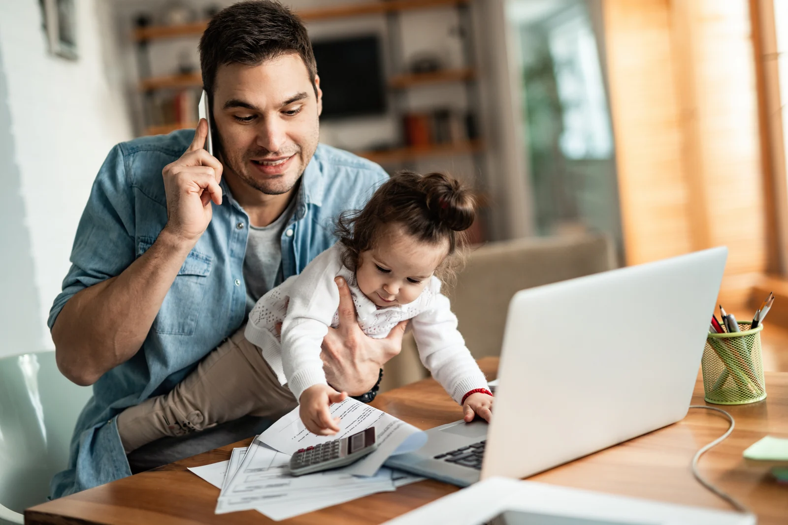Parent caring for a young child while using a phone and laptop at home.