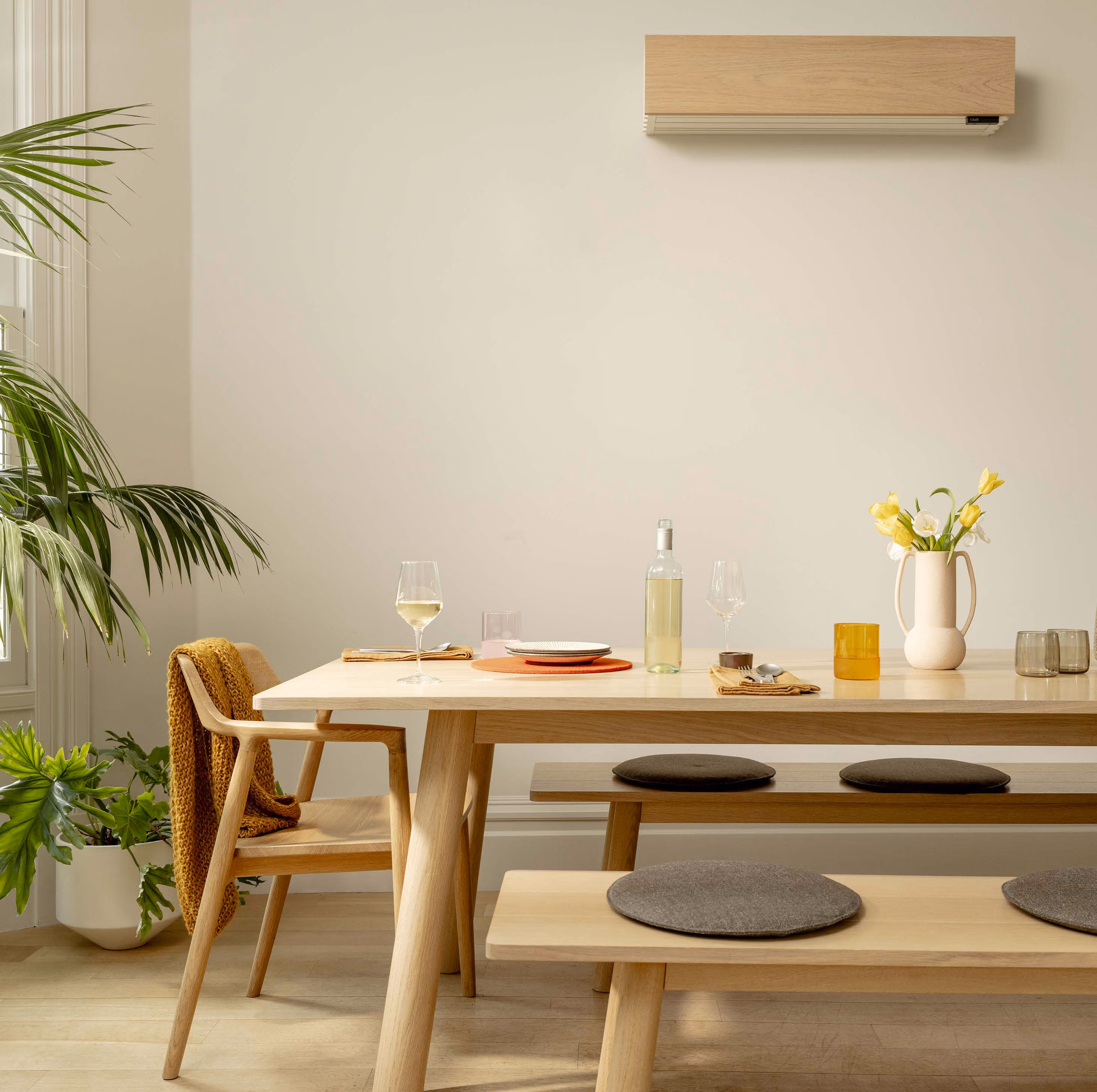 A modern dining room with a wooden table, chairs, and benches with gray cushions, decorated with a white vase holding yellow and white flowers, a bottle of white wine, wine glasses, and napkins, with green plants near a window and a wooden air conditioner on the wall.